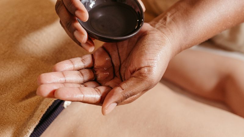 A therapist's hands pouring massage oil into her palm above a client's back.