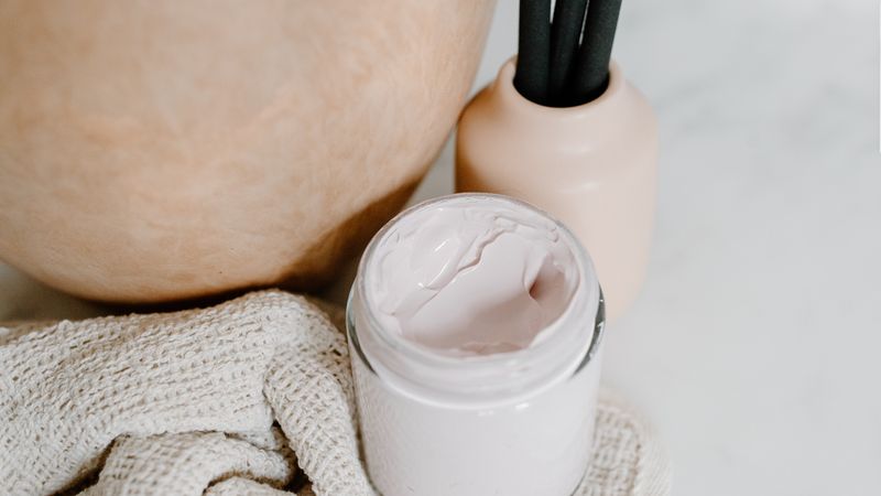An unbranded cream jar and a reed diffuser beside a folded towel, set out for a facial.