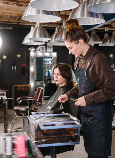 A colourist applying colour to a seated client's hair, mixing bowl in hand