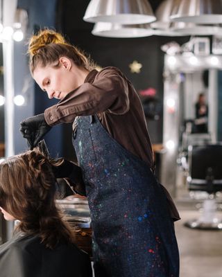 Amira Khan working on a client's hair at the mirror, wearing a paint-speckled colourist's apron